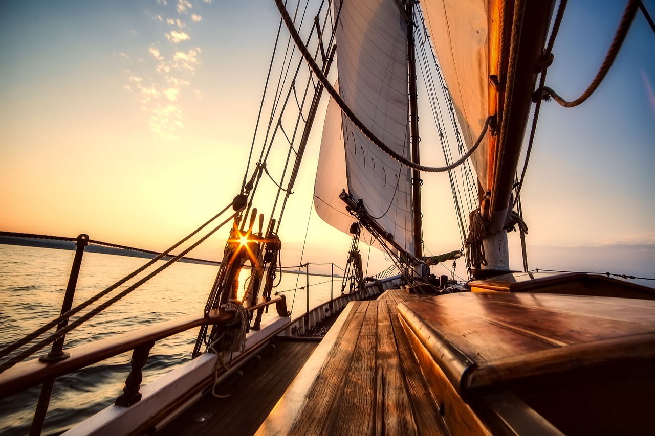 An iconic traditional American built sailing yacht on an Off-Shore Adventure, sailing into the horizon as the setting sun peaks through portside belaying pins in anticipation of the approaching sunset over a glassy sea.